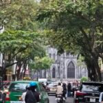 Street traffic in Hanoi with motorbikes, cars, and the cathedral visible through the trees