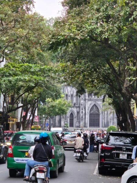 Street traffic in Hanoi with motorbikes, cars, and the cathedral visible through the trees