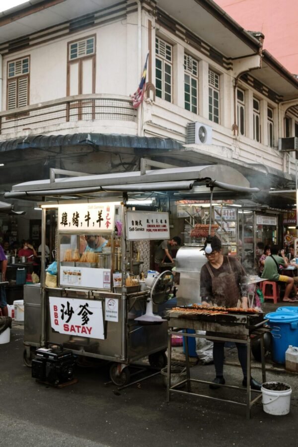 Hawker stall in George Town Penang preparing satay on a busy street with smoke and crowd