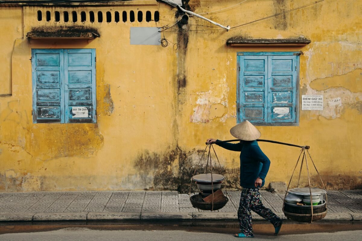 Street vendor carrying goods on a shoulder pole walking past a yellow wall with blue windows in Hoi An, Vietnam