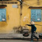 Street vendor carrying goods on a shoulder pole walking past a yellow wall with blue windows in Hoi An, Vietnam