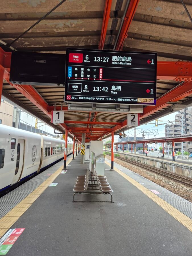 Empty platform at Futsukaichi station with a departure board showing limited express and local train times, and a train waiting on the adjacent track.