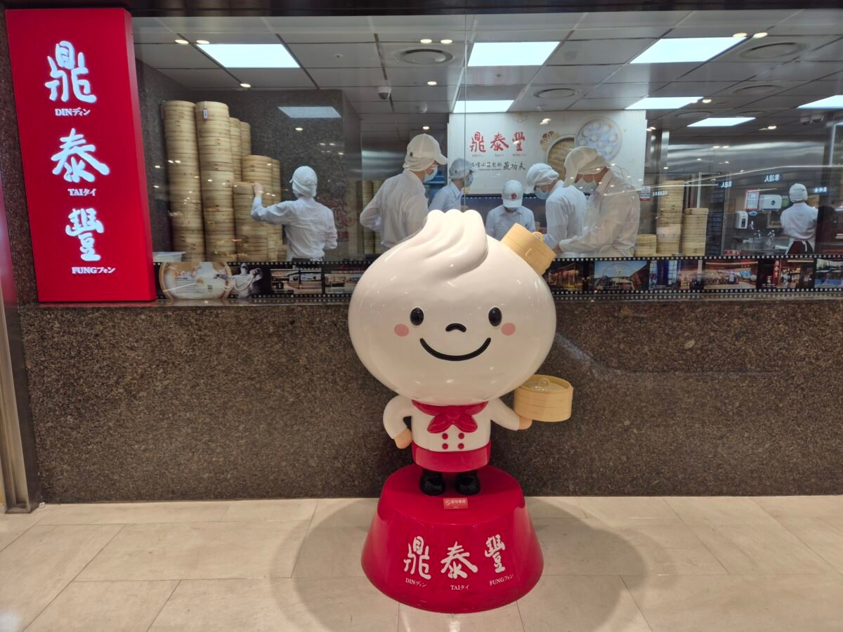 Din Tai Fung's dumpling-shaped mascot statue in chef's whites at the restaurant entrance, with staff steaming xiaolongbao in the open kitchen behind