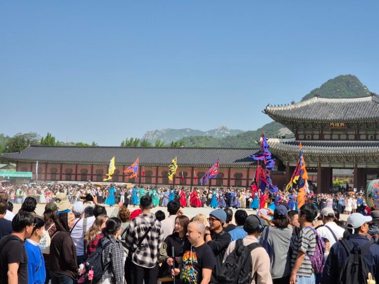Large crowd watching the royal guard changing ceremony at Gyeongbokgung Palace in Seoul, with guards in traditional dress carrying colourful flags in front of Heungnyemun gate