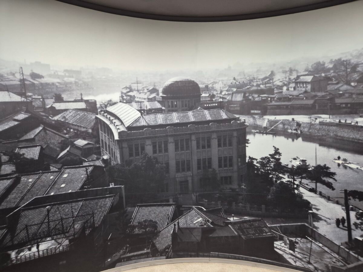 Museum display photograph of the Hiroshima Prefectural Industrial Promotion Hall, its dome intact beside the river, before the 1945 atomic bombing.