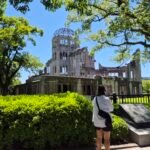 The ruined A-Bomb Dome under a clear blue sky in Hiroshima Peace Memorial Park, a visitor pausing at the memorial marker in front.