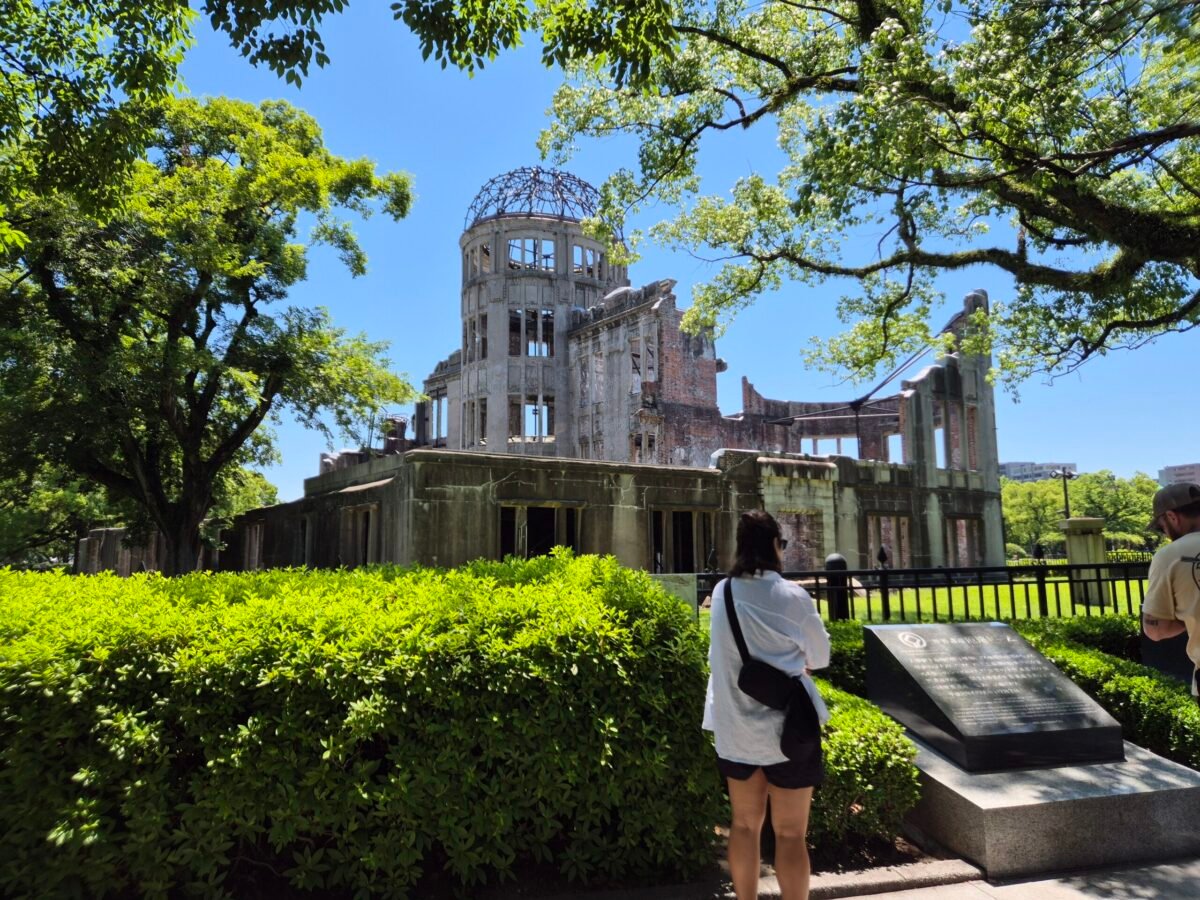 The ruined A-Bomb Dome under a clear blue sky in Hiroshima Peace Memorial Park, a visitor pausing at the memorial marker in front.