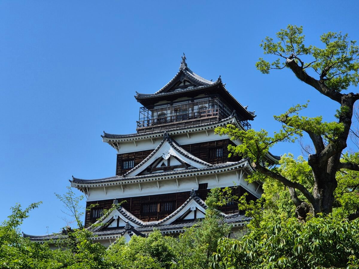 Hiroshima Castle's five-storey keep, black-and-white timber tiers against a clear blue sky, framed by green trees.