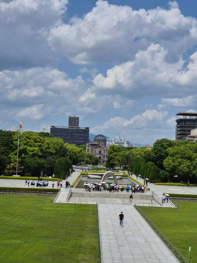 View over Hiroshima Peace Memorial Park from the museum, the Cenotaph arch, reflecting pool and A-Bomb Dome aligned beneath a cloud-filled sky.