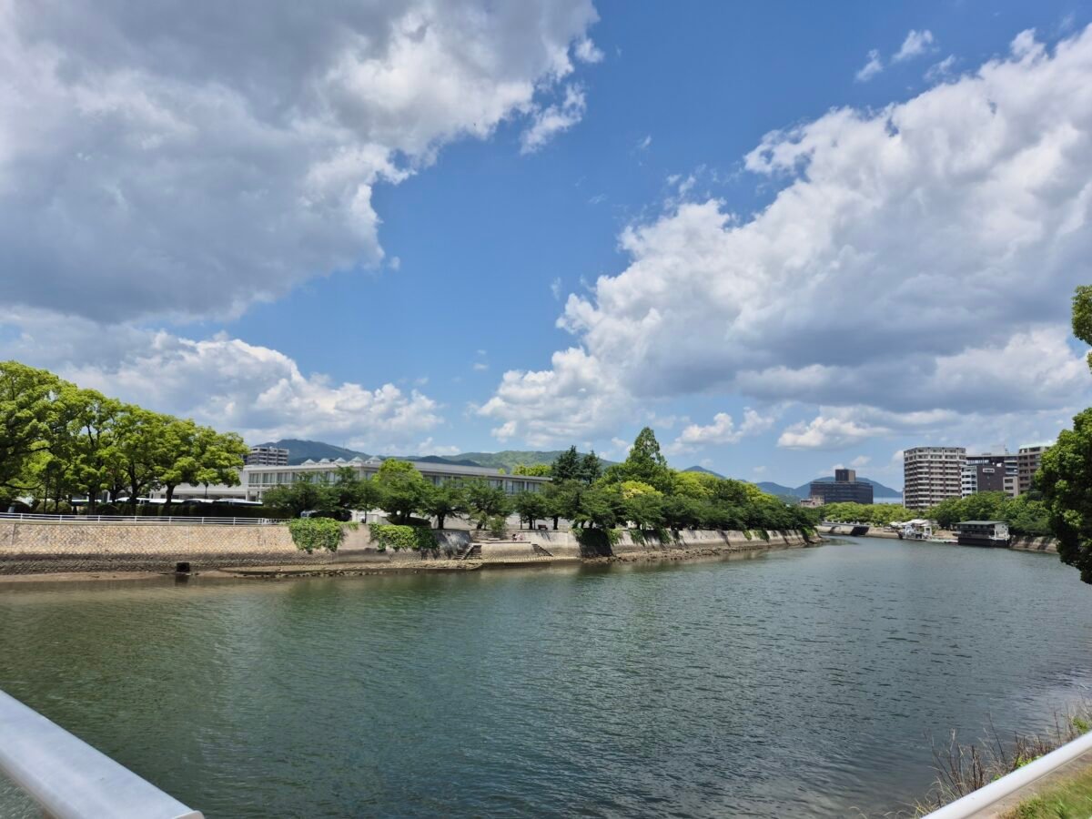 One of Hiroshima's wide rivers under a blue sky, tree-lined stone banks and green mountains beyond, the modern city along the far side.