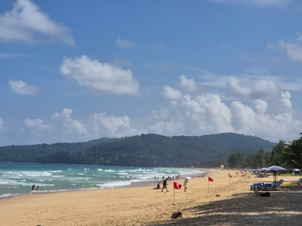 Karon Beach, Phuket, with strong waves along a long stretch of sand backed by sun loungers and green hills.