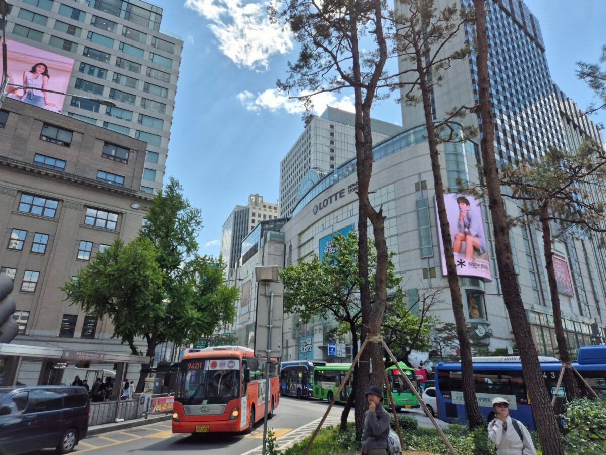 Busy Myeongdong street in Seoul near Lotte Department Store, with city buses, traffic, and pedestrians crossing beneath high-rise buildings