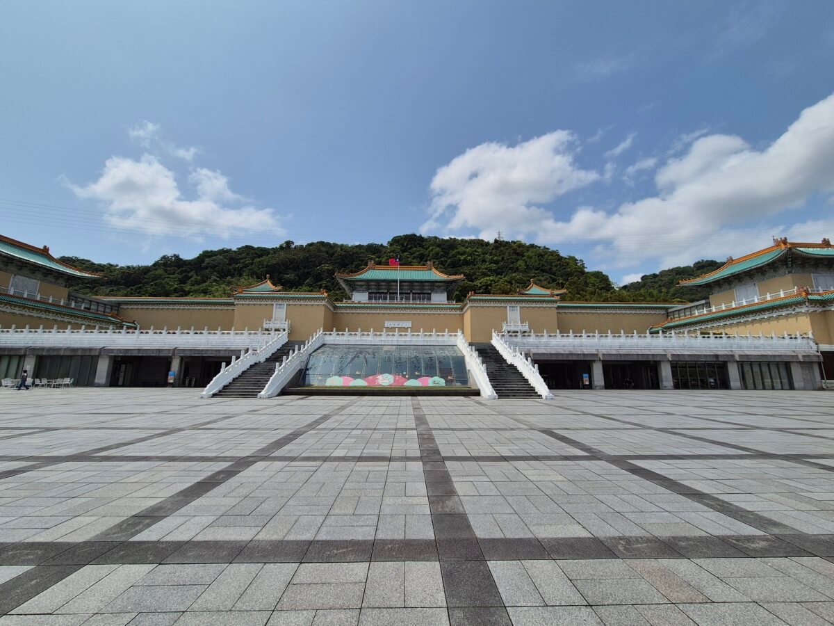 National Palace Museum in Taipei, its green-roofed pavilion and twin marble staircases framed by a forested hill, seen across the empty front plaza