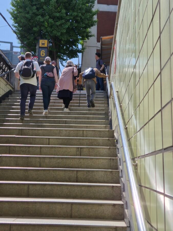 Long uncovered staircase at Exit 6 of a Seoul subway station, with no lift and travellers carrying backpacks climbing to street level