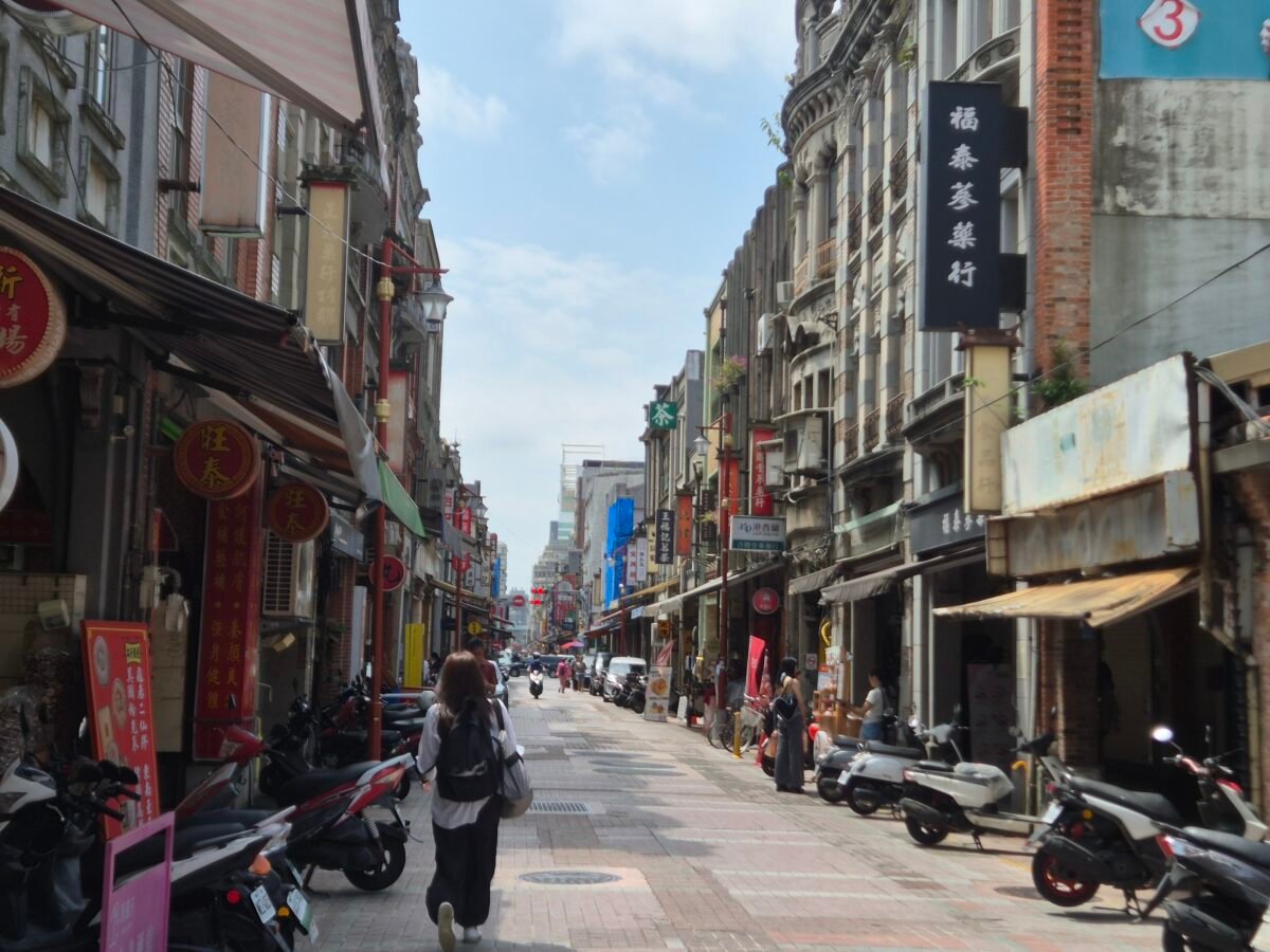A bustling old Taipei street lined with ornate baroque-style shophouses, herbal medicine shop signs, parked scooters, and pedestrians