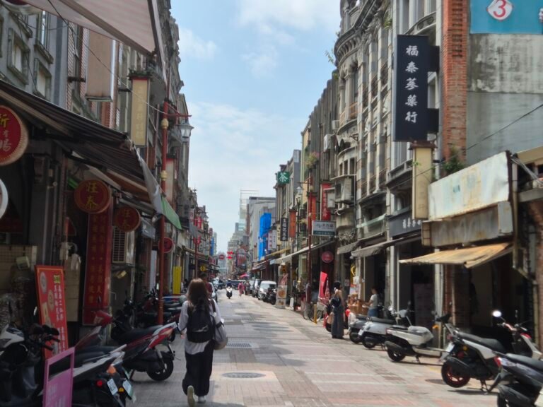 A bustling old Taipei street lined with ornate baroque-style shophouses, herbal medicine shop signs, parked scooters, and pedestrians